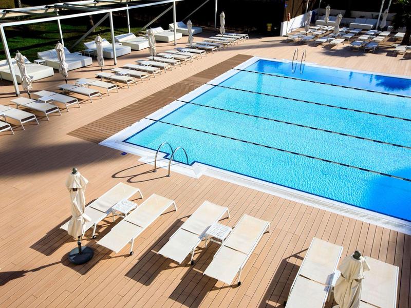 Gran piscina al aire libre con tumbonas blancas y sombrillas en una terraza de madera bajo el sol.