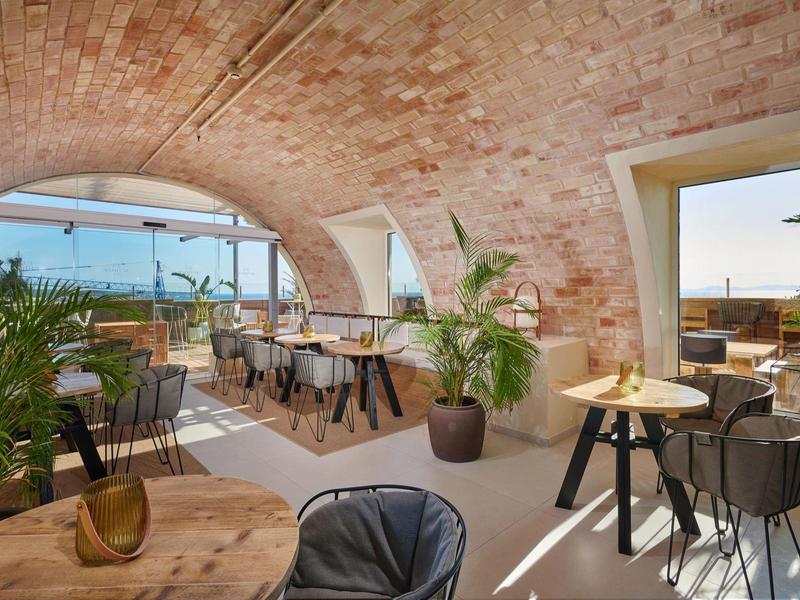 Restaurante luminoso con techo abovedado de ladrillo, mesas de madera y plantas con vista a la ciudad.