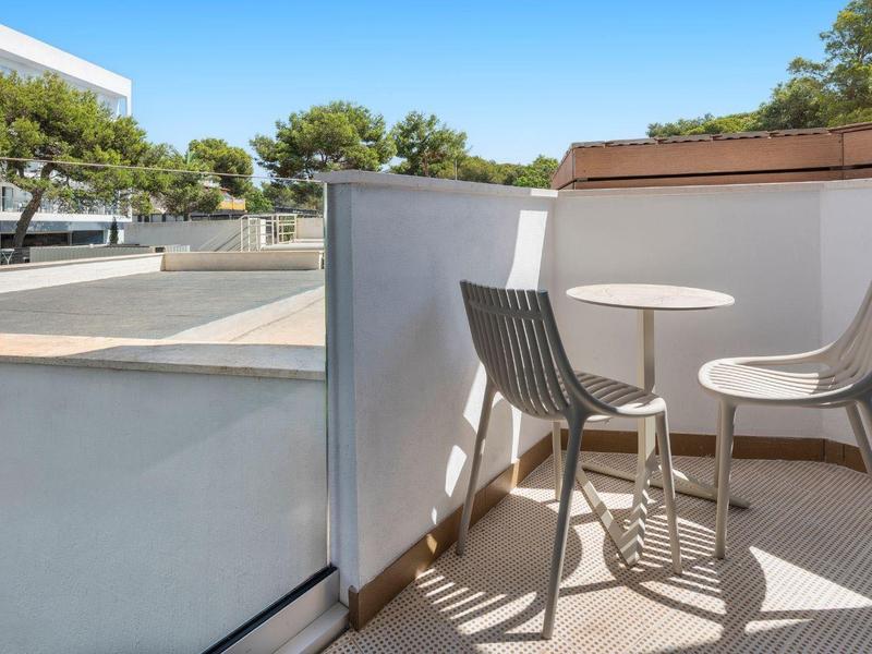 Balcón con dos sillas y mesa redonda pequeña, edificios y árboles visibles alrededor.