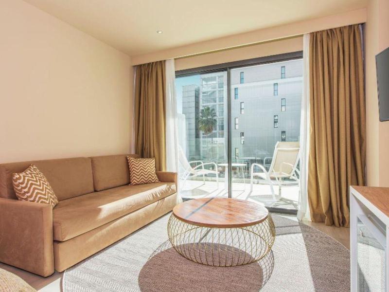 Habitación de hotel moderna con sofá marrón, dos mesas redondas y acceso a terraza con sillas.