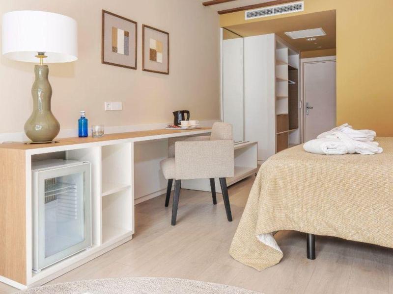 Habitación de hotel luminosa con escritorio, silla, minibar, cama y paredes amarillas cálidas.