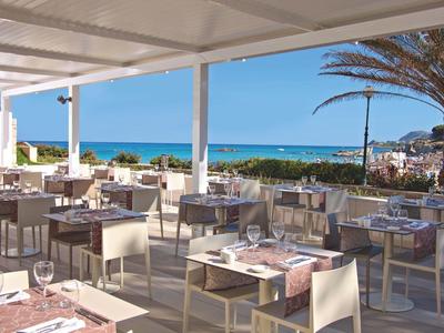 Offenes Restaurant mit weißen Tischen und Stühlen, Blick auf Meer und Palmen unter blauem Himmel.