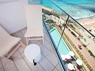 Balkon mit weißen Stühlen und rundem Tisch neben Glasgeländer mit Blick auf belebten Strand.