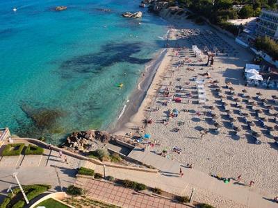 Klares Meer, Sandstrand mit Sonnenliegen und Pool im Vordergrund, bewaldete Hügel im Hintergrund.