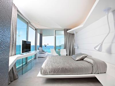 Helles Schlafzimmer mit großen Glasfenstern, grauem Bett und Meerblick im Hintergrund.