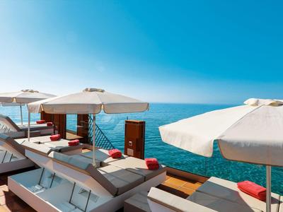 Sonnige Dachterrasse mit weißen Liegen, großen Sonnenschirmen und Blick auf das blaue Meer.