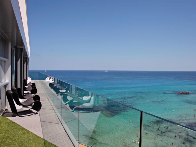 Balkon mit Glasgeländer und Stühlen neben klarem, türkisblauem Meer unter klarem Himmel.