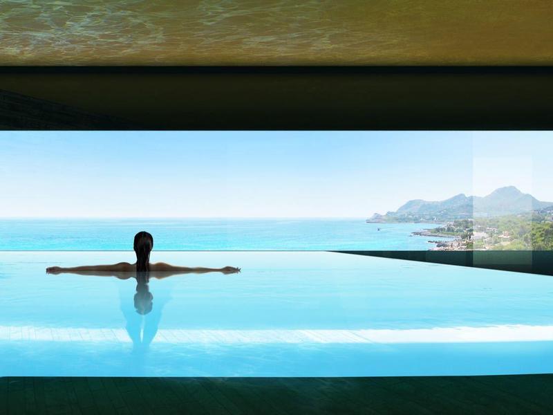 Person entspannt in großem Infinity-Pool mit Blick auf Meer, Berge und blauem Himmel.
