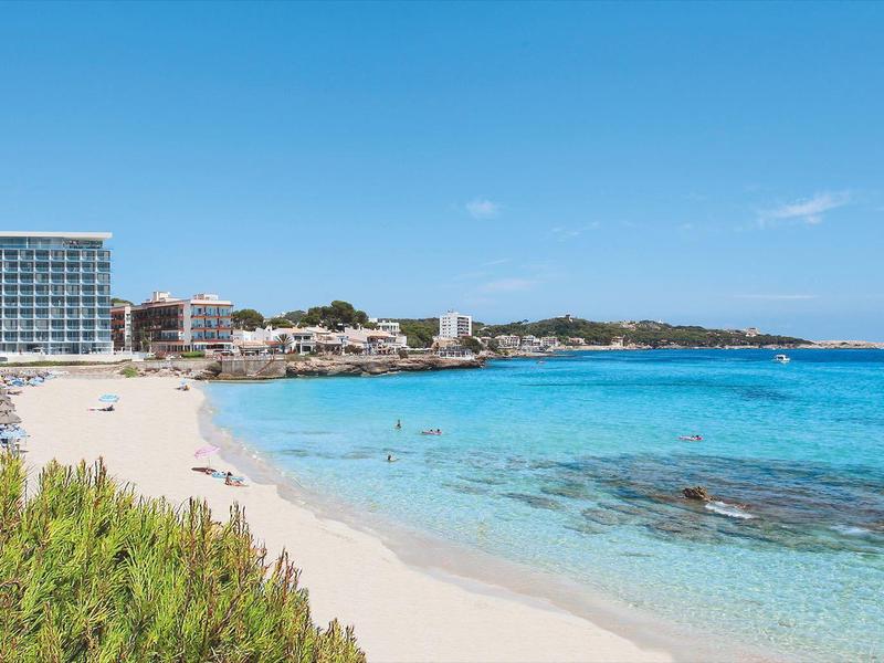 Strand mit klarem blauem Wasser, feinem Sand und Gebäuden am Ufer unter blauem Himmel.