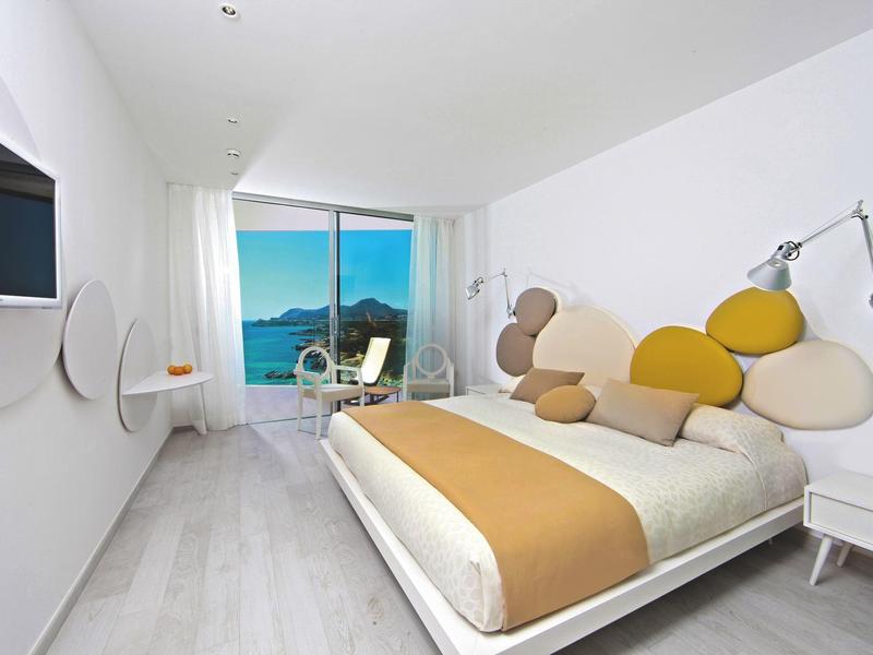 Modernes Schlafzimmer mit weißem Bett, gelb-beigem Bettbezug und großem Fenster mit Meerblick.