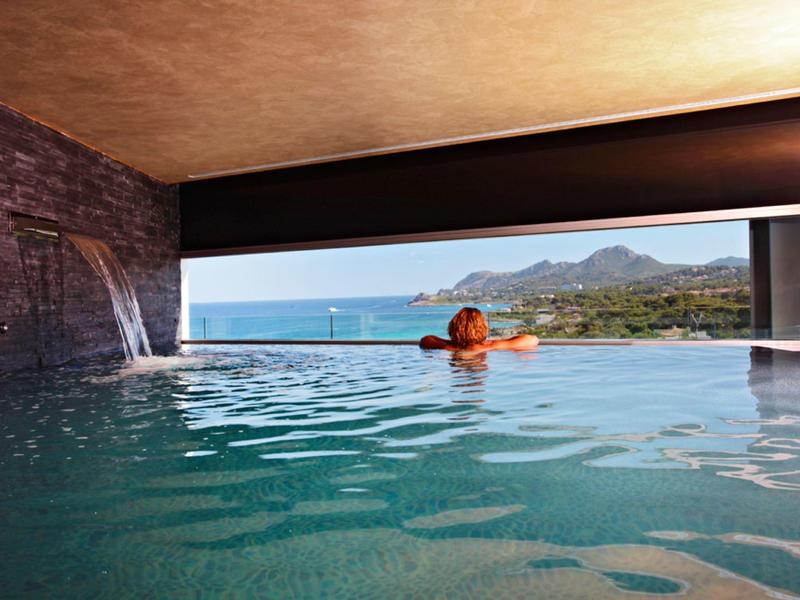 Frau im Infinity-Pool blickt auf Meer und Berge bei klarem Himmel, holzverkleidete Decke, Wasserfall.