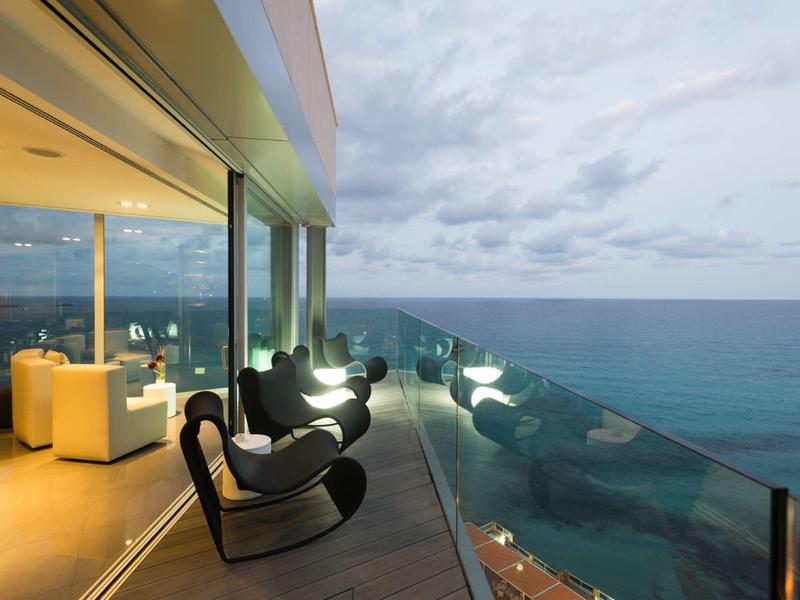 Moderne Terrasse mit schwarzen Schaukelstühlen, Glaskante und Blick auf das Meer bei bewölktem Himmel.