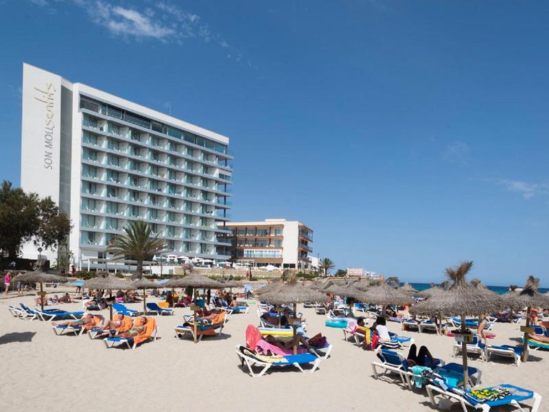 Strand mit Sonnenschirmen, Liegestühlen und mehreren Urlaubern vor einem mehrstöckigen Hotel.