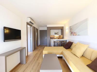 Salon lumineux avec canapé jaune, table basse, TV murale et sol en bois dans une chambre d'hôtel.