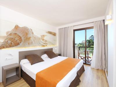 Pohodlný hotelový pokoj s manželskou postelí, oranžovým přehozem a balkonem s výhledem na zeleň.