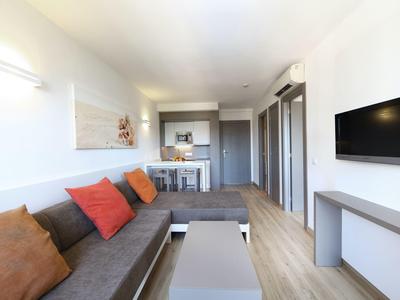 Moderní obývací pokoj se šedou pohovkou, barevnými polštáři, televizí na zdi a balkonem v pozadí.