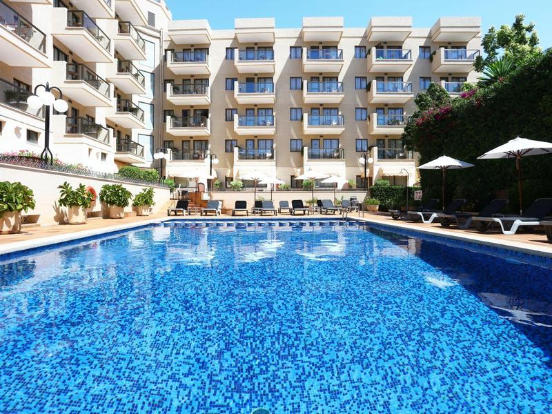 Hotelový bazén s lehátky před moderní budovou pod jasnou oblohou.