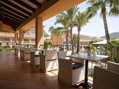 Offene Terrasse mit Tischen und Stühlen, palmen und Hotelgebäude im Hintergrund.
