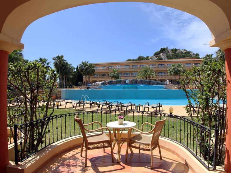 Terrasse mit Tisch und zwei Stühlen mit Blick auf Pool und Hotelgebäude unter blauem Himmel.