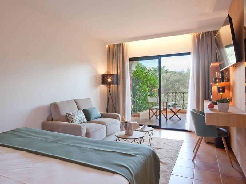 Helles Zimmer mit Bett, Sofa, Tisch, Fernseher und Balkon mit Blick auf grüne Landschaft.