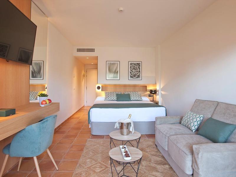 Helles Hotelzimmer mit Doppelbett, Sofa, Schreibtisch, zwei Tischen und Wandbildern.