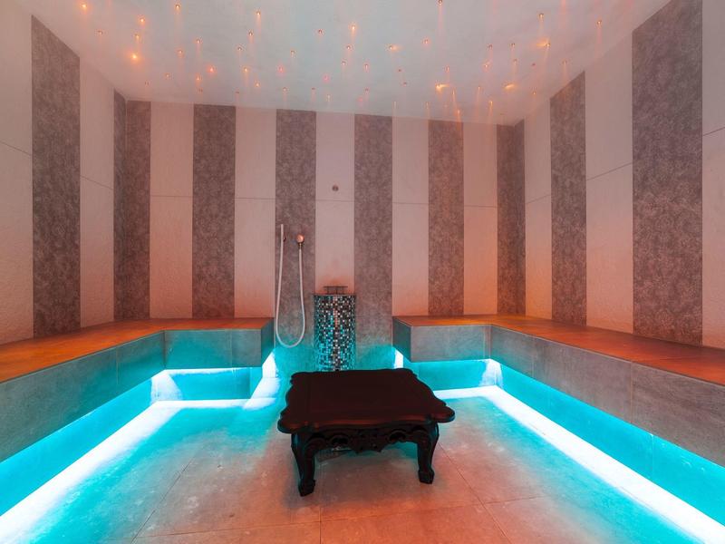 Sala de spa moderna con piscina iluminada, paredes de mosaico y mesa baja en el centro.