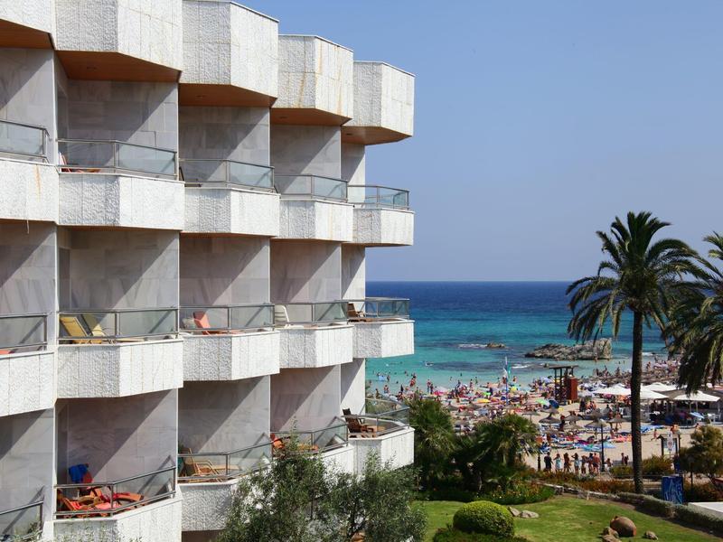 Hotel con balcones, vista a la playa y el mar bajo cielo despejado con palmeras.