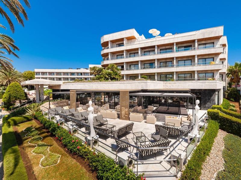 Hotel moderno con terraza al aire libre, sillas y mesas, rodeado de jardines y cielo azul.