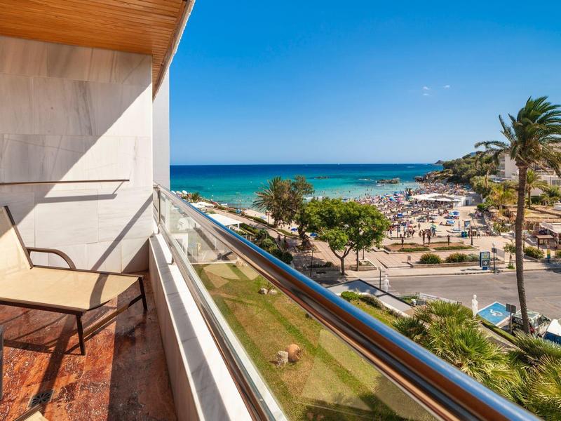 Balcón con sillas y vista al mar y playa en un hotel soleado y tropical.