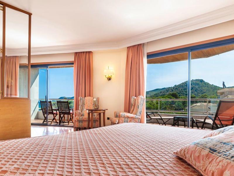 Habitación de hotel espaciosa con cama doble, balcón y vista panorámica de colinas verdes de día.