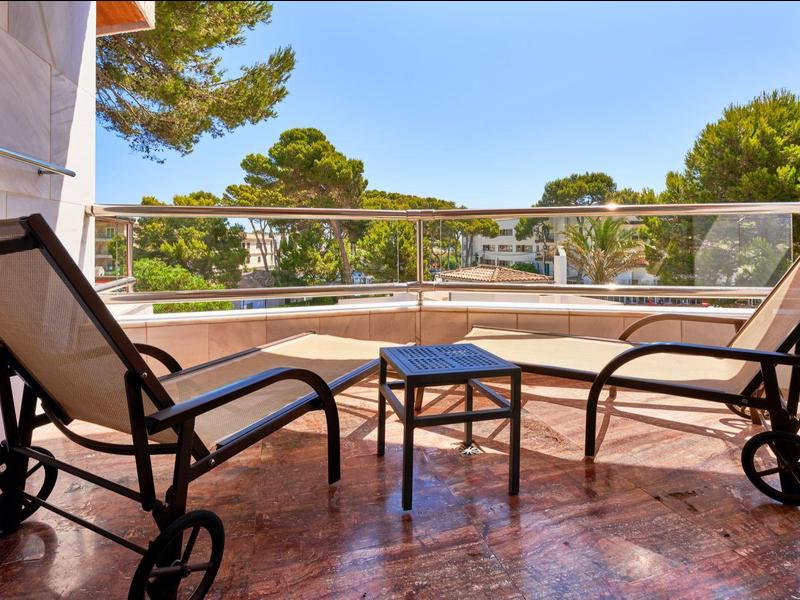 Balcón con dos tumbonas y una mesa pequeña, rodeado de árboles y cielo azul.