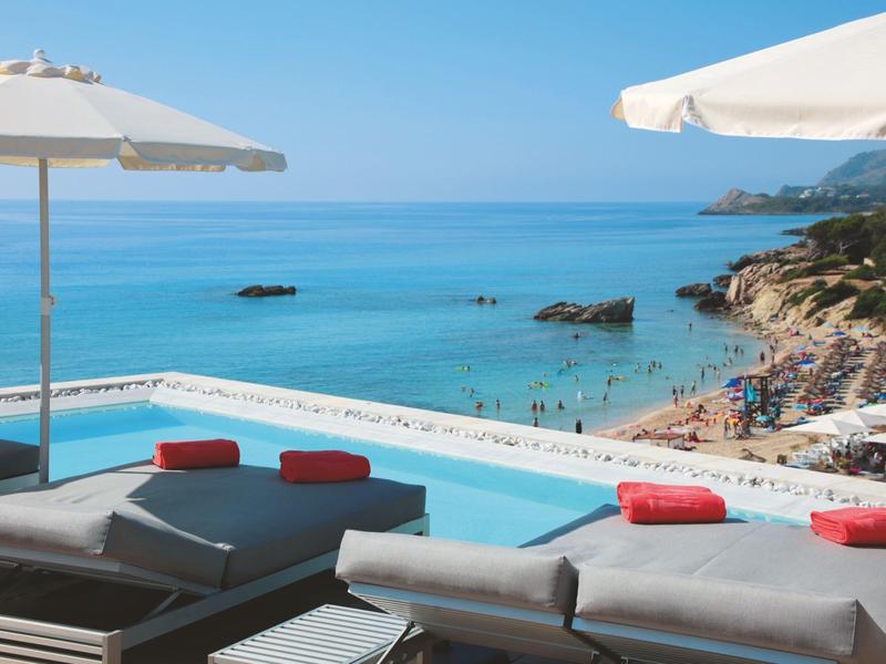 Piscina con tumbonas y sombrillas con vista a la playa y al mar azul.