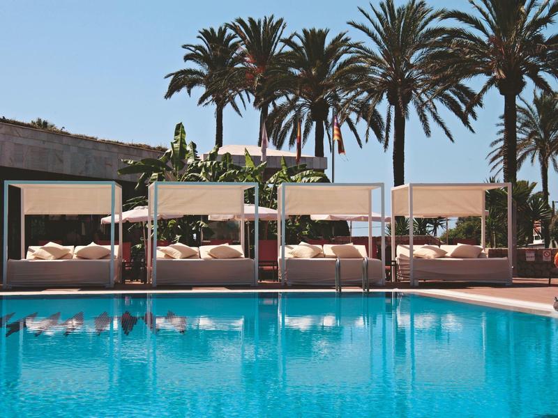 Piscina del hotel con agua azul, cuatro camas balinesas y palmeras al fondo en un día soleado.