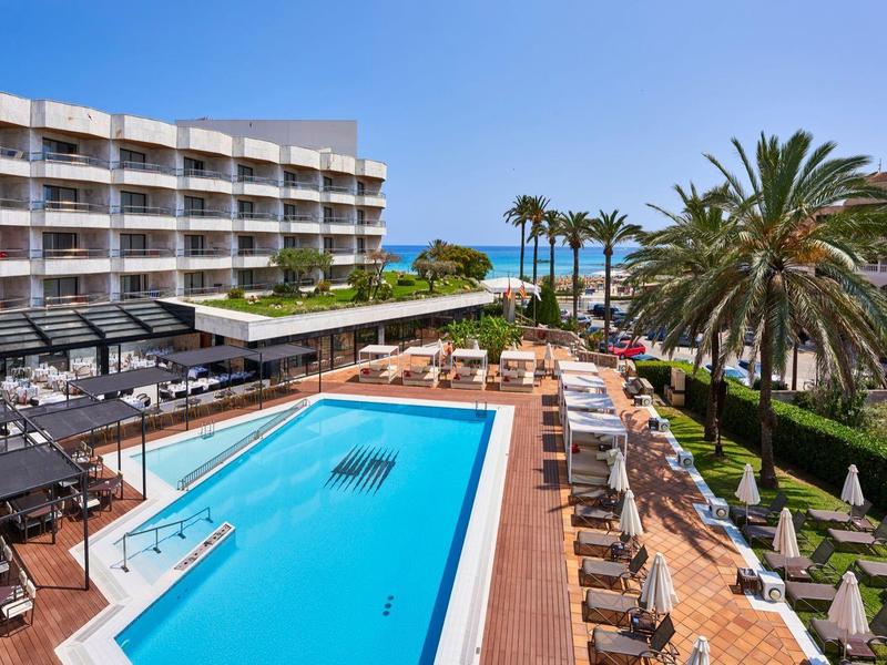 Hotel con gran piscina, tumbonas y palmeras, vista al mar al fondo.
