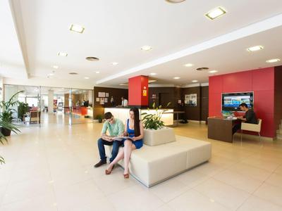 Bright hotel lobby with seating, two women relaxing, and check-in area in the background.