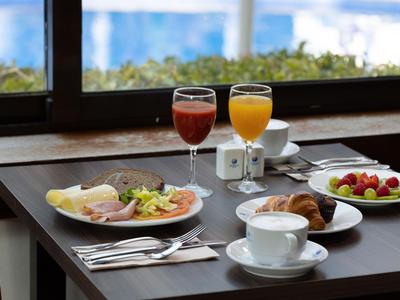 Breakfast table with coffee, croissant, meats, fruit, and juices by window with plants outside.