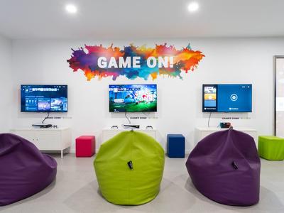 Sala giochi moderna con tre schermi, pouf colorati e scritta "GAME ON!" sul muro.