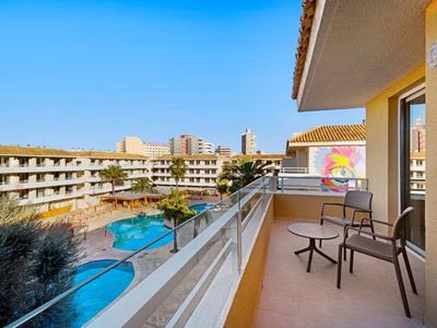 Balkon mit Stühlen und Tisch, Blick auf Pool, Palmen und mehrstöckiges Hotel unter blauem Himmel.
