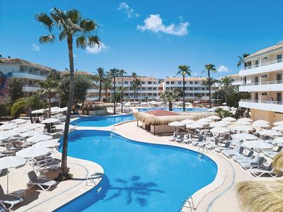 Großer blauer Pool mit weißen Liegestühlen und Palmen vor mehrstöckigem Hotel bei Sonnenschein.