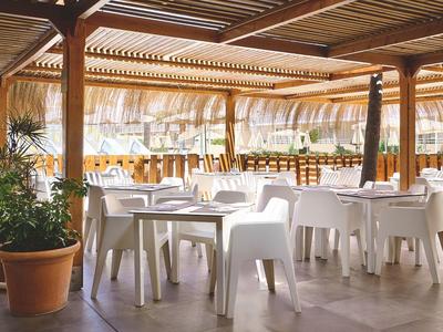 Hell beleuchtete Terrasse mit weißen Stühlen und Tischen unter Holzdach, dekoriert mit grünen Pflanzen.