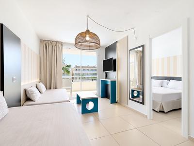 Helles Wohnzimmer mit Sofa, blauem Tisch, TV und Blick ins angrenzende Schlafzimmer.