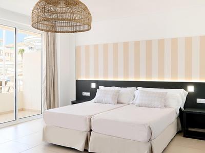 Hell eingerichtetes Schlafzimmer mit zwei weißen Betten, braunem Lampenschirm und Glas-Schiebetür.