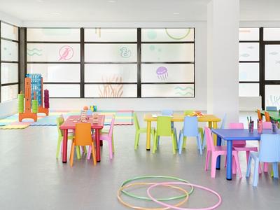 Heller Raum mit bunten Kindertischen, Stühlen und Spielgeräten, großen Fenstern und grauem Boden.
