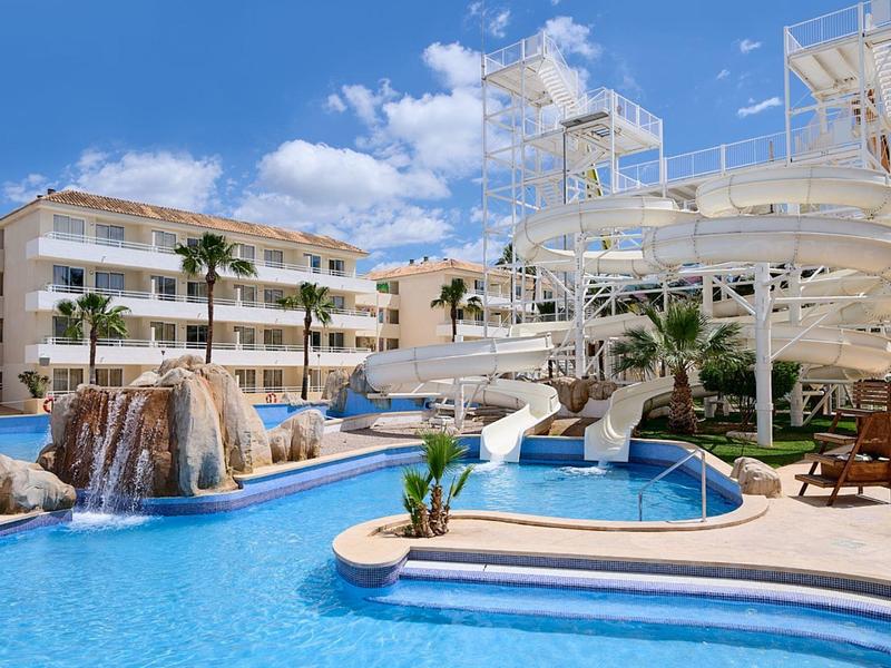 Piscina d'hotel con scivoli d'acqua, cascate, palme e sdraio sotto un cielo azzurro.