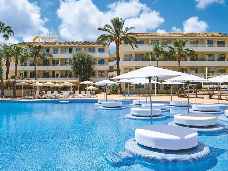 Hotel moderno con piscina e grandi ombrelloni bianchi sotto un cielo azzurro.