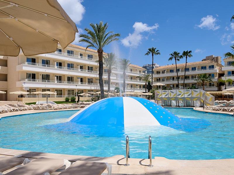 Piscina dell'hotel con ombrelloni, palme e edificio sullo sfondo sotto cielo blu.