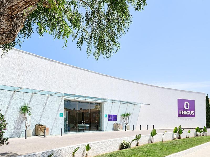 Edificio bianco moderno con insegna viola, ingresso vetrato, alberi e prato verde.