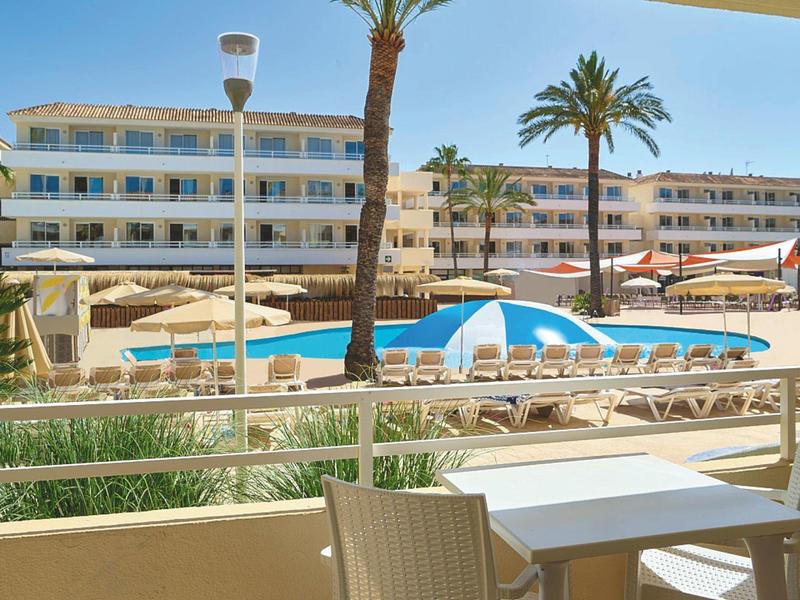 Hotel con piscina all'aperto, lettini, ombrelloni e palme sotto un cielo sereno.