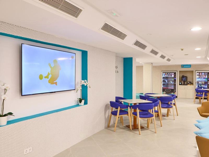 Moderner Raum mit blau-gelben Stühlen, Tischen, großem Bildschirm und heller minimalistischer Einrichtung.