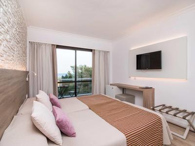 Bright hotel room with double bed, TV, and balcony overlooking trees.